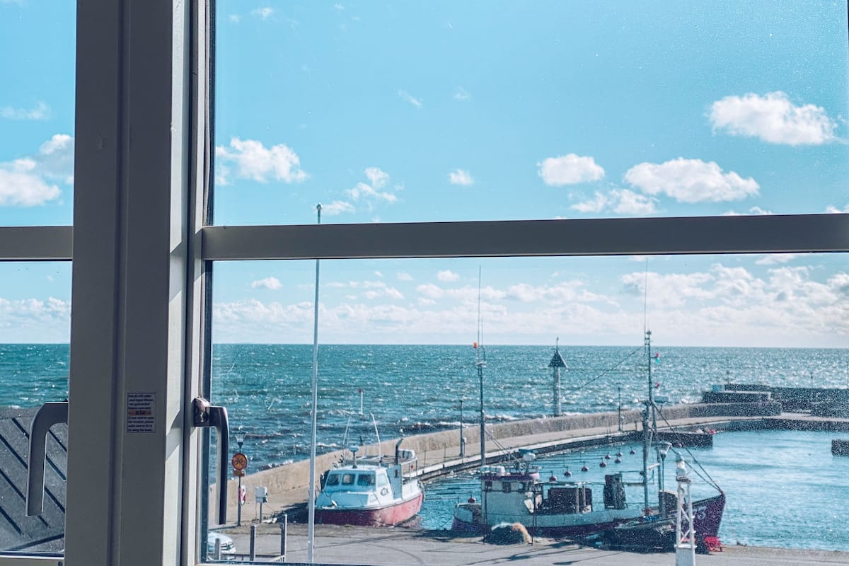 Vista a través de una ventana de un puerto con barcos amarrados, cielo despejado y mar en calma.
