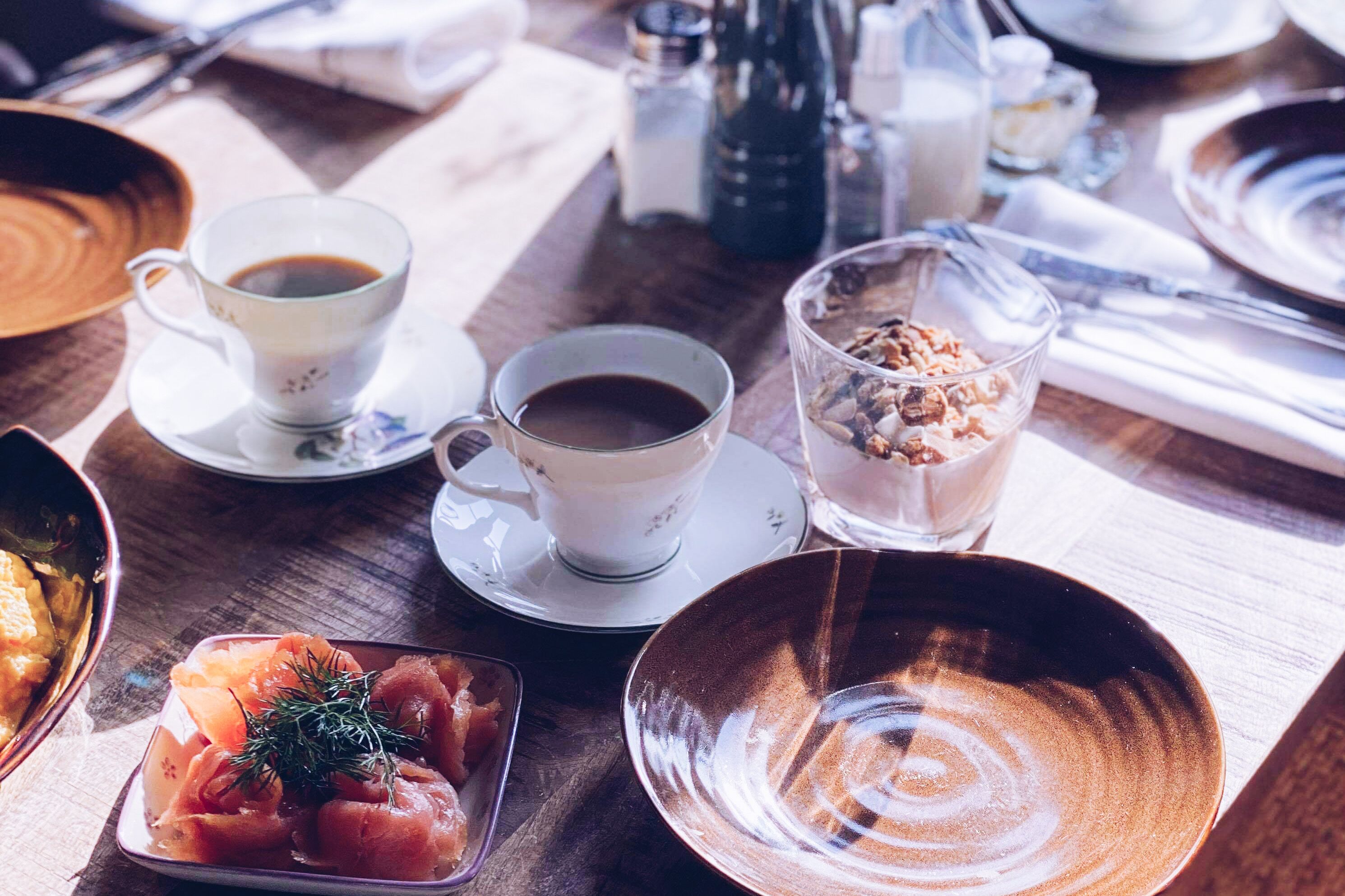 Ein Frühstückstisch mit Kaffeetassen, einem Glas Granola und Teller mit Speisen auf einer Holztischplatte.