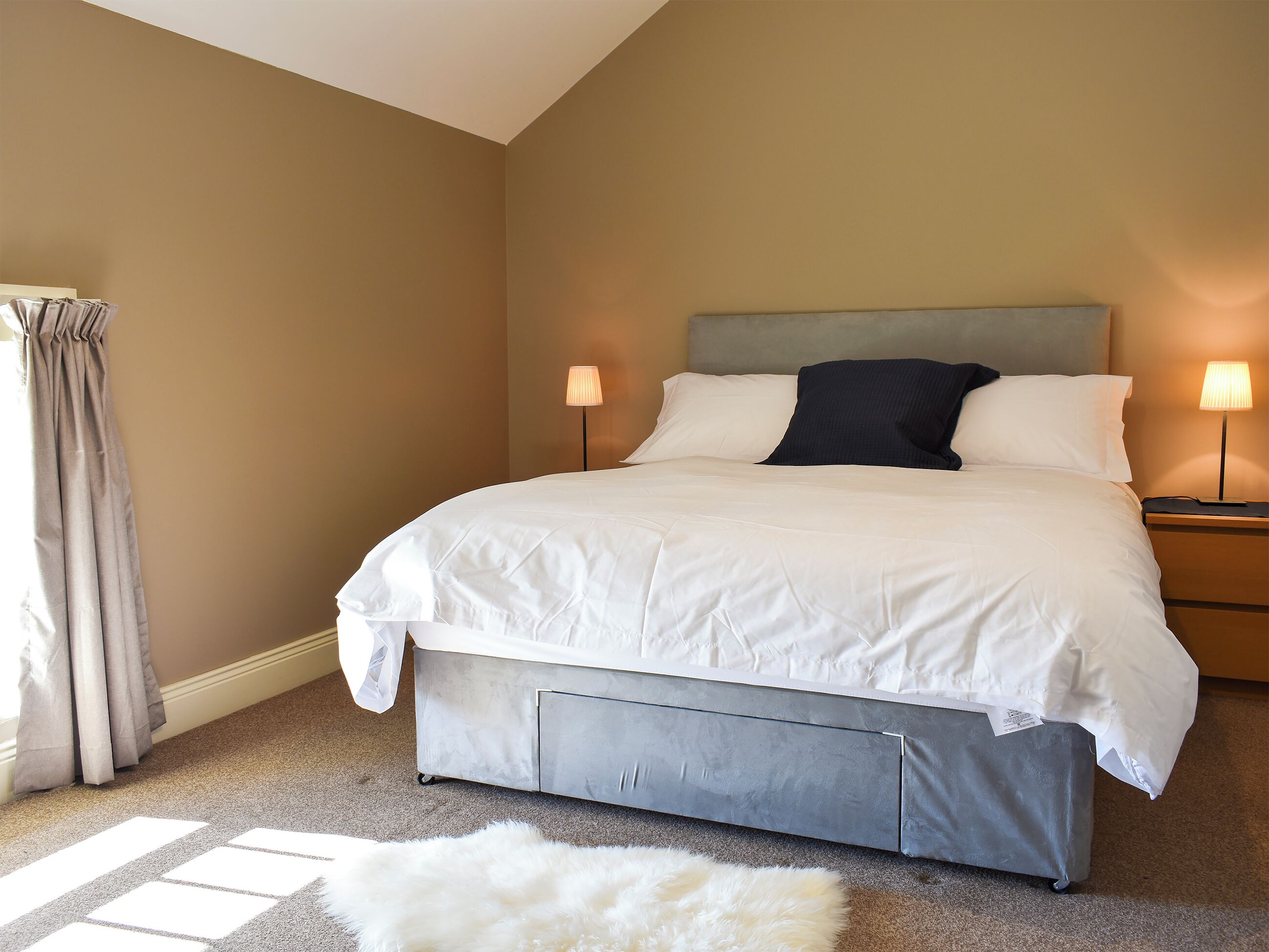 A neatly made bed with a grey headboard, two bedside lamps, and a fluffy white rug on a carpeted floor.