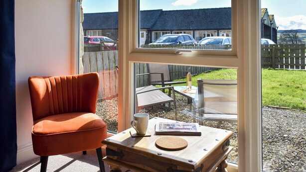 A room with a large window, a wooden table, a red chair, and a view of parked cars and houses outside.