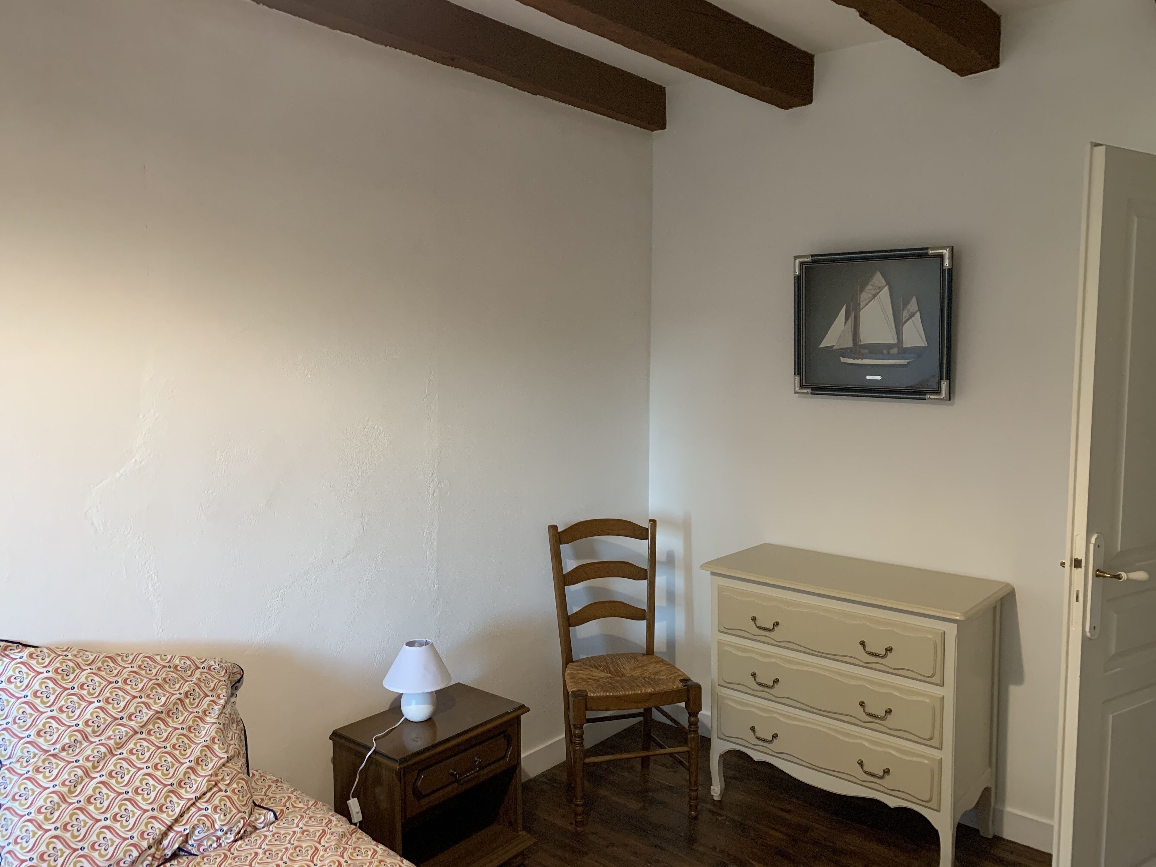 A bedroom with a bed, a nightstand with a lamp, a wooden chair, a dresser, and a framed picture on the wall.