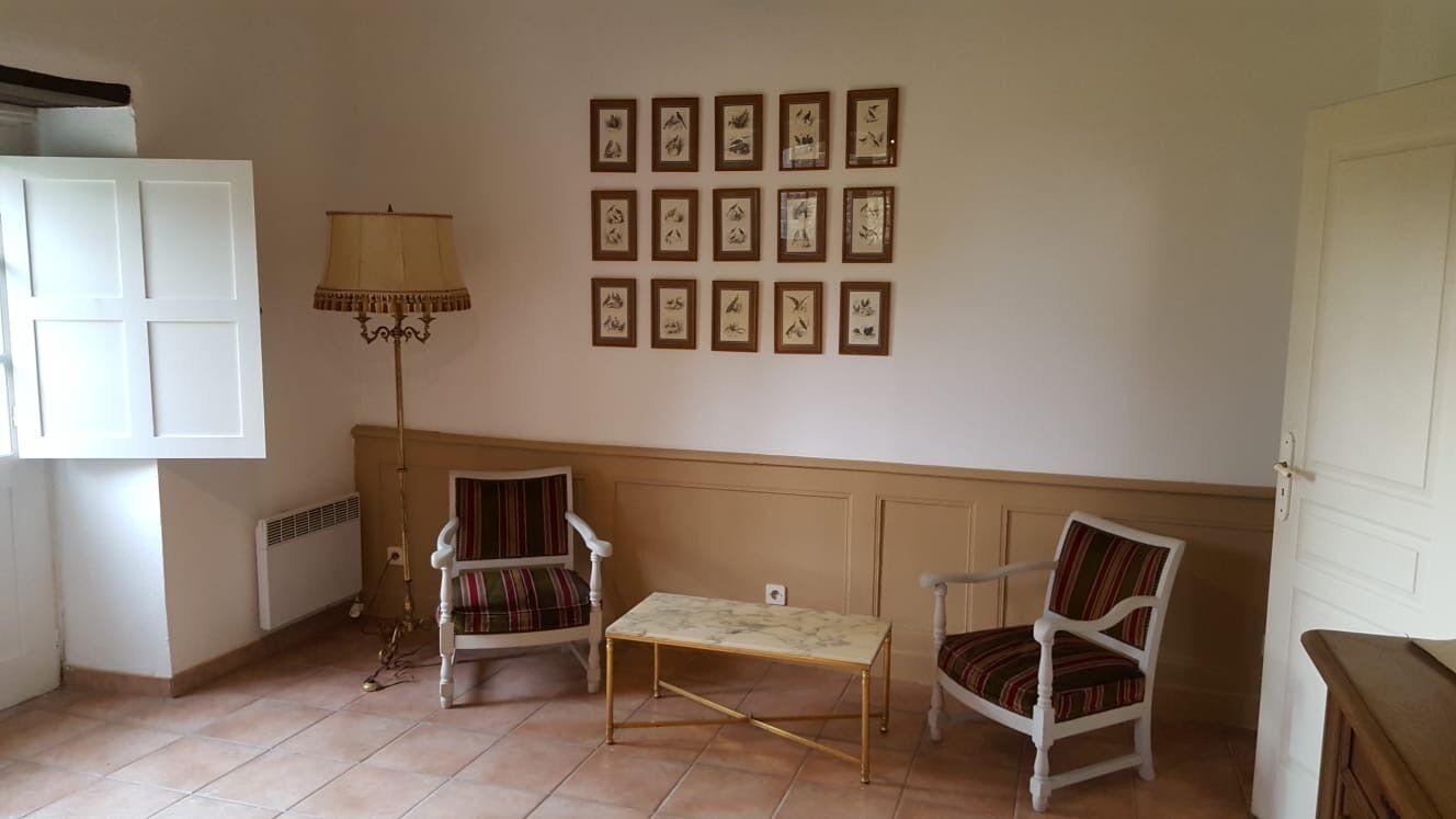 Une pièce avec un meuble blanc, un lampadaire, deux fauteuils, une petite table et un mur orné de tableaux encadrés.