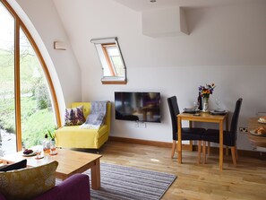Uma sala de estar moderna com um sofá amarelo, uma mesa de jantar com cadeiras, uma televisão de tela plana na parede e uma grande janela com vista para o verde.