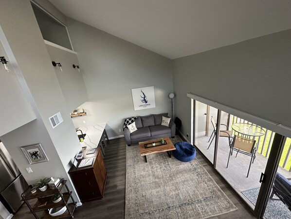 Una sala moderna con un sofá gris, una mesa de centro y un sillón de peluche azul. Hay una zona de comedor con una mesa y sillas de madera. Un ventanal ofrece vistas al exterior.