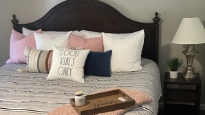 Een netjes opgemaakt bed met gestreept beddengoed, meerdere kussens, een dienblad met een potje vanille, en een kleine potplant op een nachtkastje.