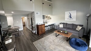 Een ruime woonkamer met een grijze bank, een salontafel en een blauwe poef. Achtergrond is een keuken met een wit aanrecht en barstoelen. Op de muur hangt een lijst met een afbeelding van Lake Conroe, Texas.