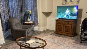 Un salon doté d’un grand téléviseur à écran plat, d’un fauteuil gris, d’un canapé à motifs, d’une table basse en verre et d’un tableau représentant une carte du monde accroché au mur.