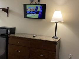 Uma área de cozinha com micro-ondas, um pequeno controle remoto, uma TV fixada na parede exibindo um anúncio e um abajur.