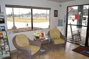 Quarto com duas poltronas, uma mesinha com um vaso, uma janela com vista para um estacionamento e uma geladeira.