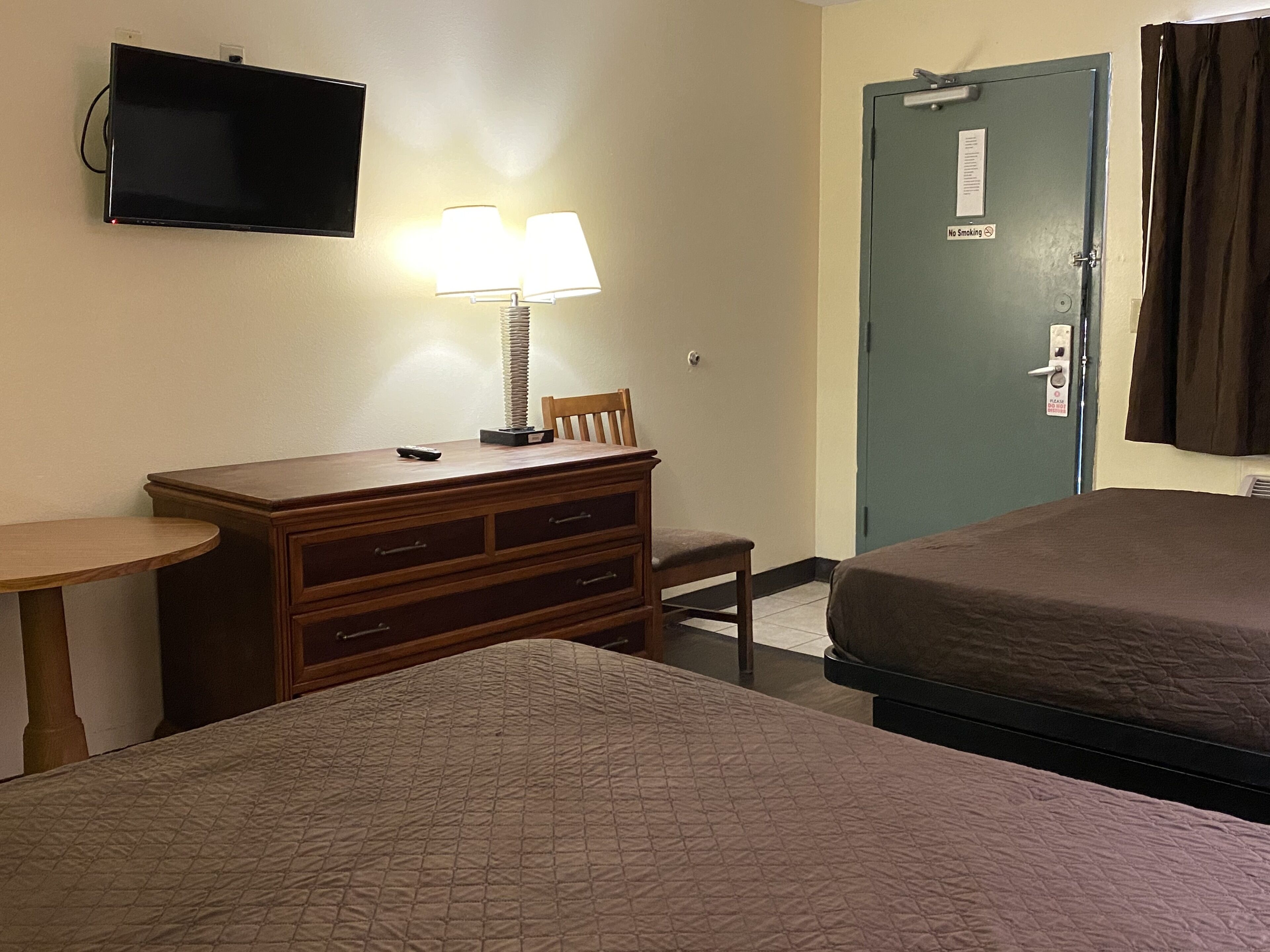 Une chambre d’hôtel avec un lit, un bureau doté d’une lampe, un téléviseur fixé au mur et une porte sur laquelle est apposé un panneau « Interdiction de fumer ».