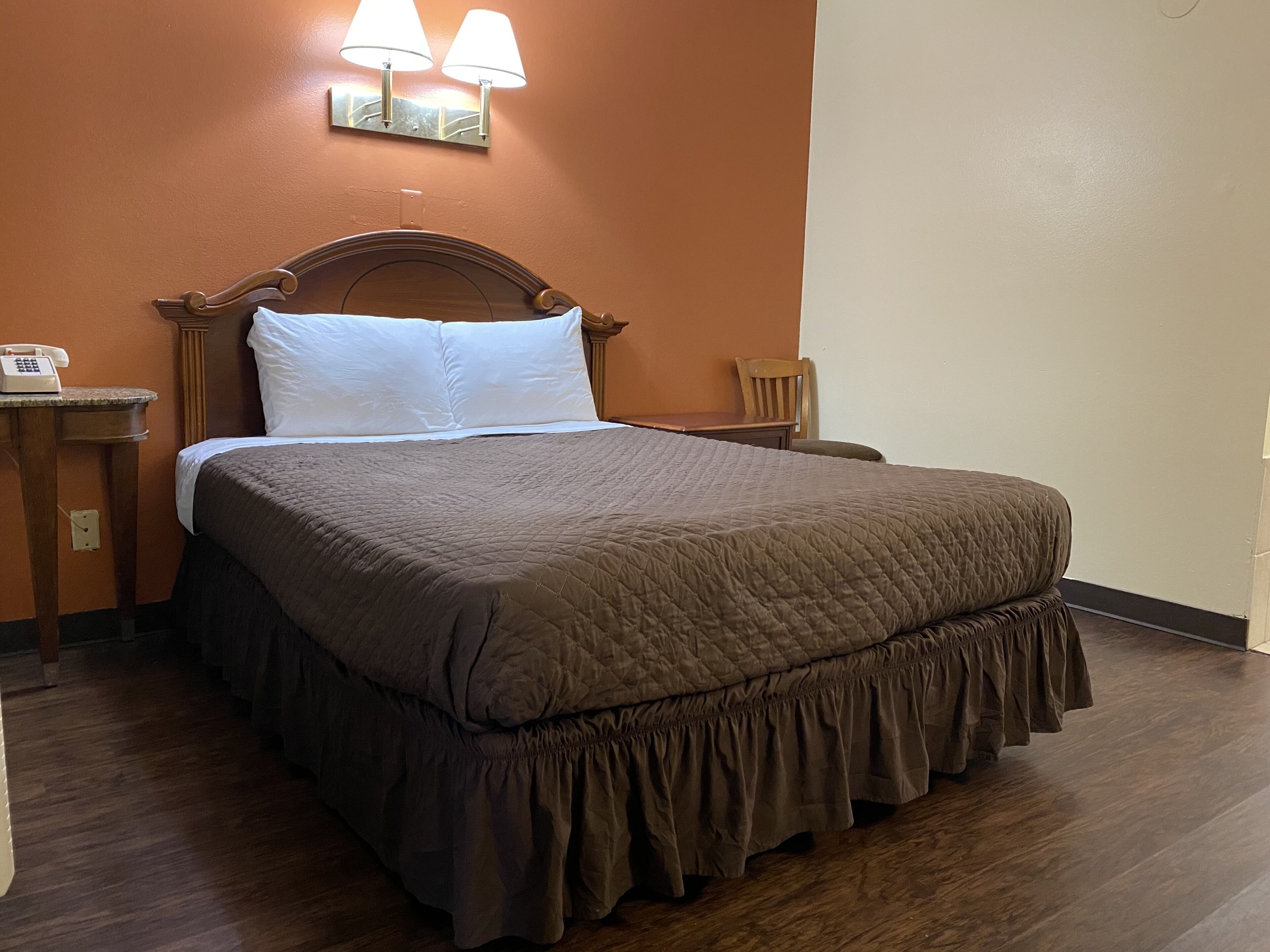 Una cama bien tendida con una colcha marrón y almohadas blancas, una mesita de noche de madera con un teléfono, y una lámpara fijada a la pared.