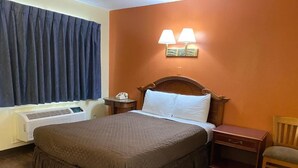A hotel room with a large bed, a bedside table, a chair, a wall-mounted lamp, and a window with curtains.