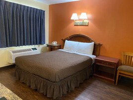 Quarto de hotel com uma cama grande, uma mesinha de cabeceira, uma cadeira, uma luminária na parede e uma janela com cortinas.
