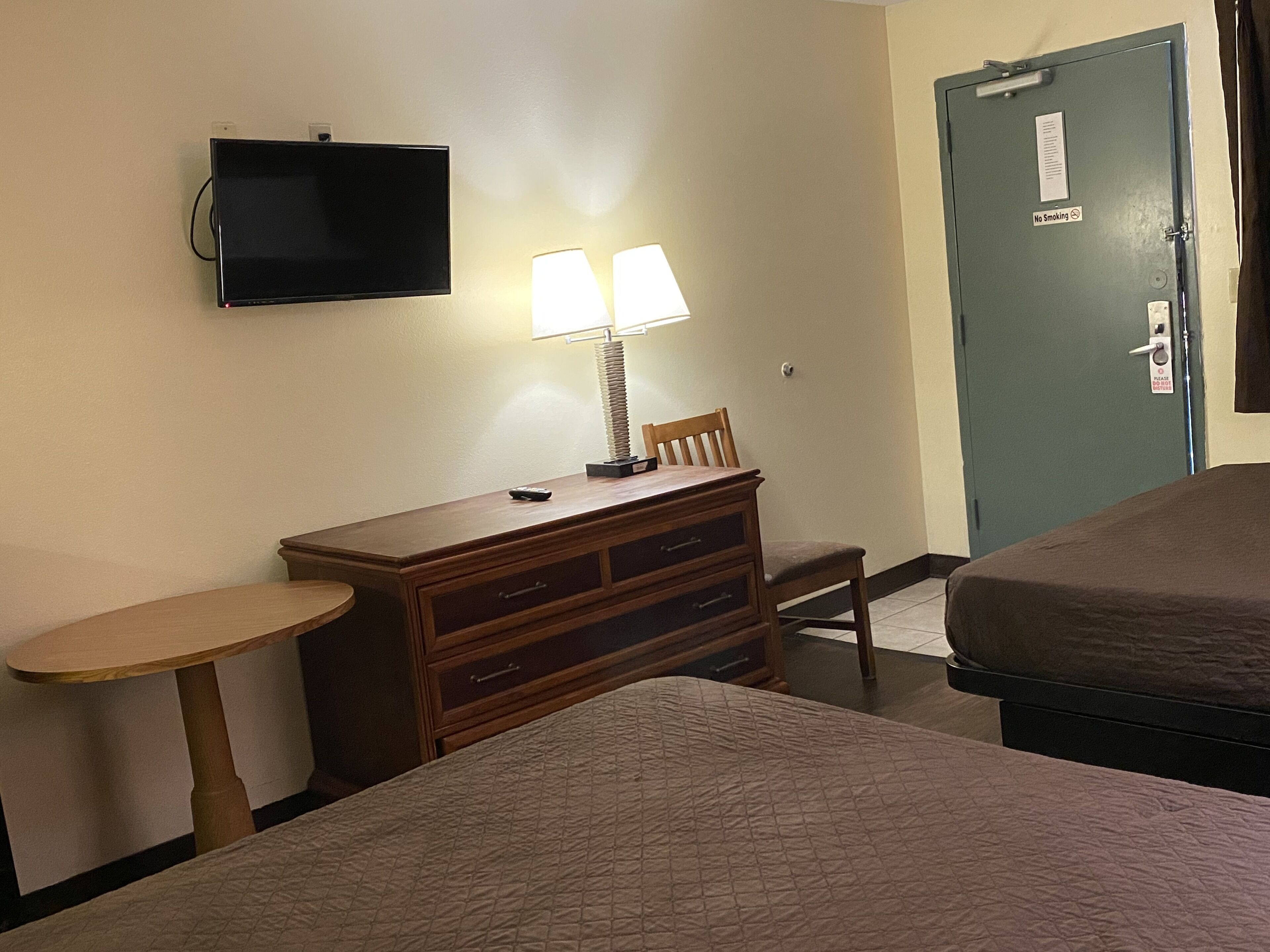 A hotel room with a bed, a desk with a lamp, a television, and a door with a "No Smoking" sign.