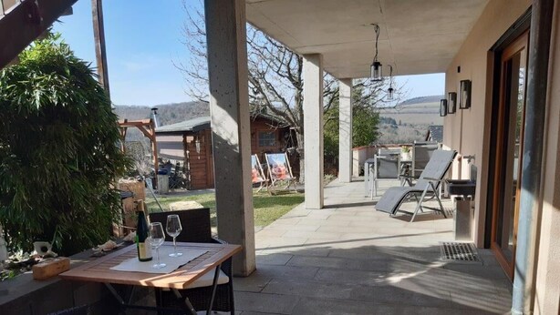 A covered patio with a table set for two, a chair, and a bottle of wine. - Large vacation apartment in the beautiful Ruwer valley (Kasel)