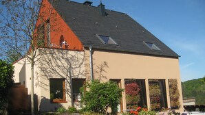 A two-story house with a sloped roof, a balcony on the upper floor, and a garden with various plants and flowers. - Large vacation apartment in the beautiful Ruwer valley (Kasel)