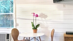 Une pièce moderne avec une table ronde, deux chaises, une orchidée en pot, un téléviseur fixé au mur et une fenêtre avec des stores.