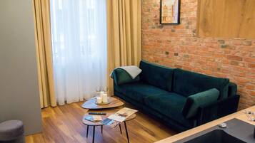 A cozy living room with a brick wall, a green sofa, a small round table with a remote control and magazines, and a window with sheer curtains.