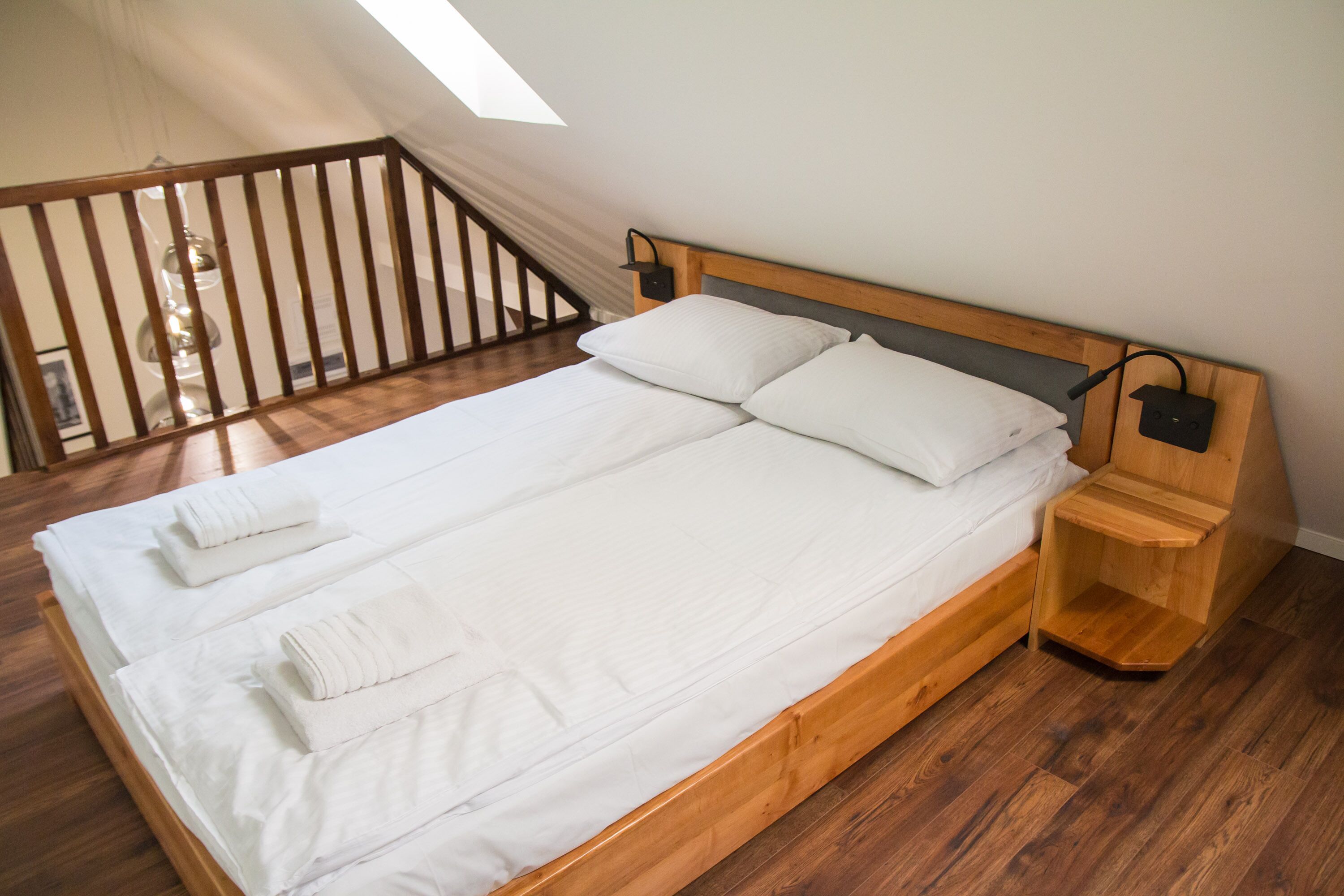 A double bed with white linens and two towels, a wooden headboard, a bedside table with a lamp, and a wooden floor.