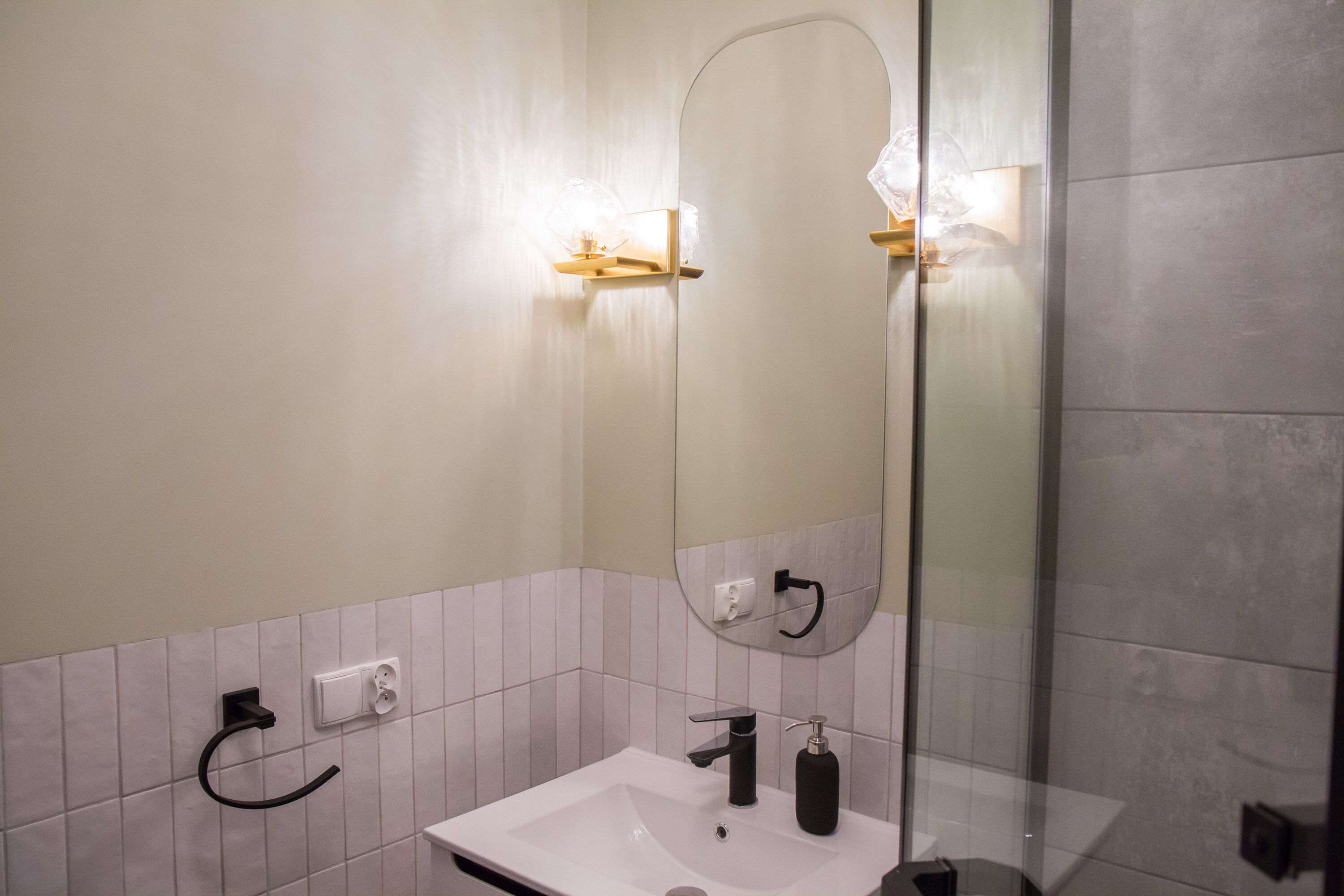 A bathroom with a white sink, a black faucet, and a mirror. There is a towel rack and a soap dispenser on the wall.