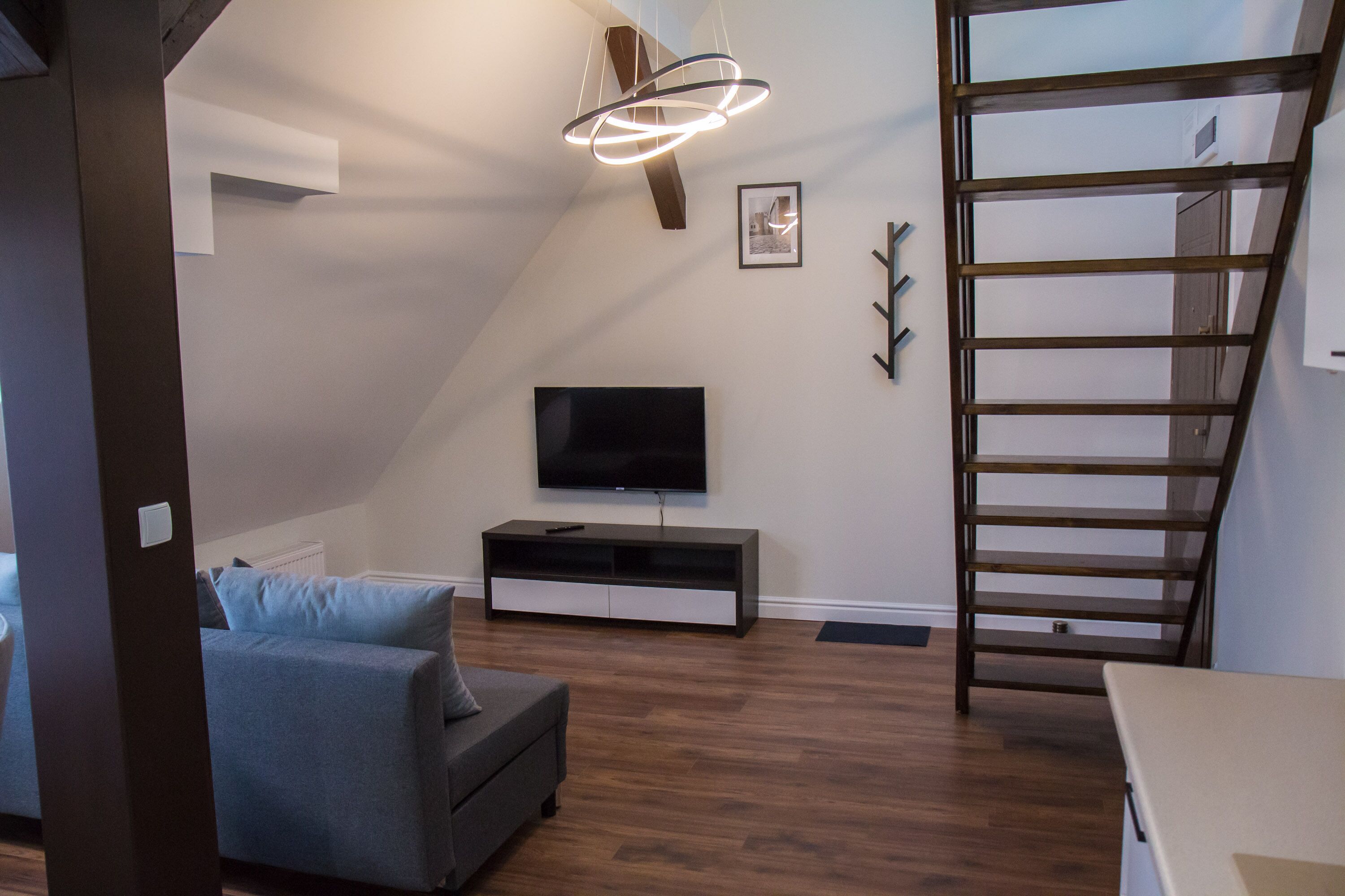 A modern living room with a flat-screen TV, a grey sofa, wooden flooring, a ladder, and a ceiling light fixture.