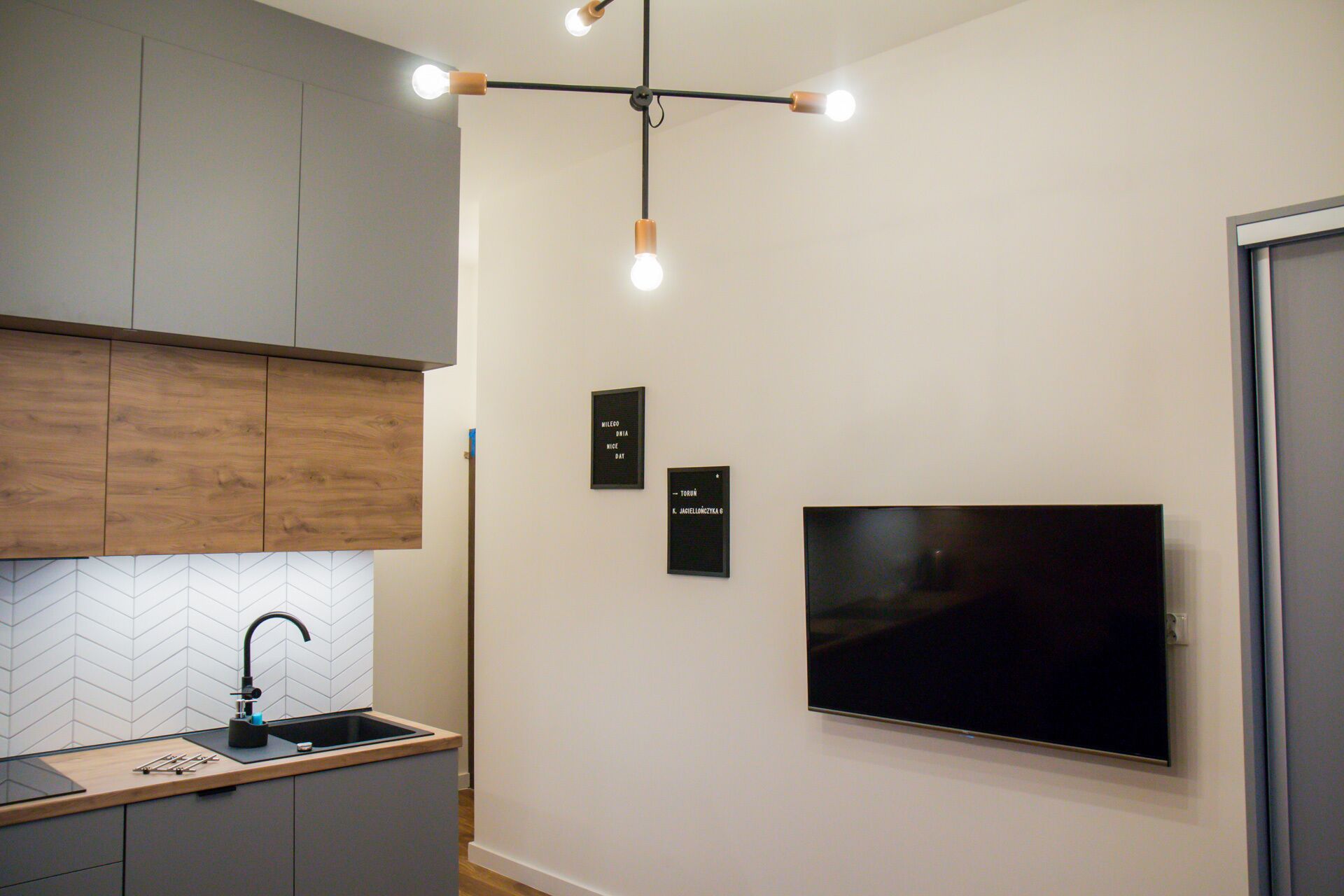 A modern kitchen with a built-in sink, wooden cabinets, a herringbone-patterned backsplash, and a flat-screen TV mounted on the wall.
