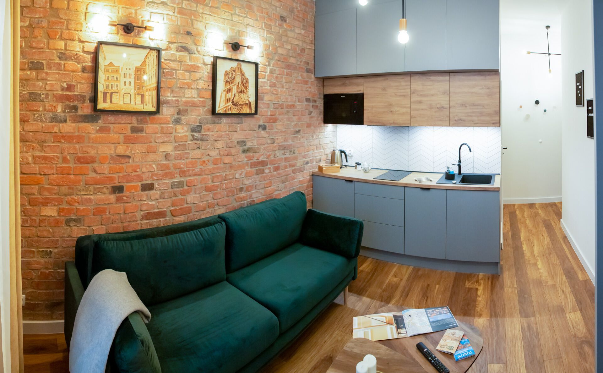 A compact living space with a green sofa, a kitchen area with grey cabinets, and a brick wall with two framed pictures.