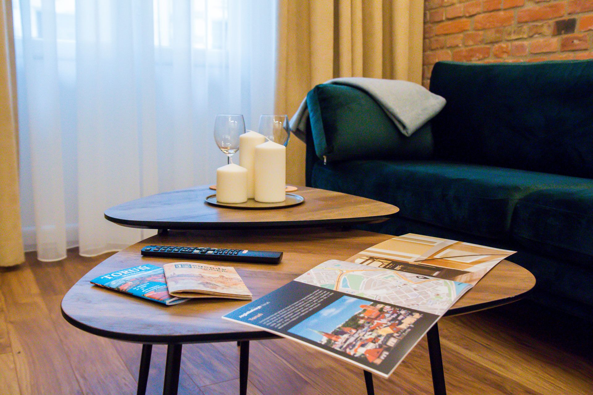A small round table with a remote control, a newspaper, and a brochure. There is a green sofa and a brick wall in the background.