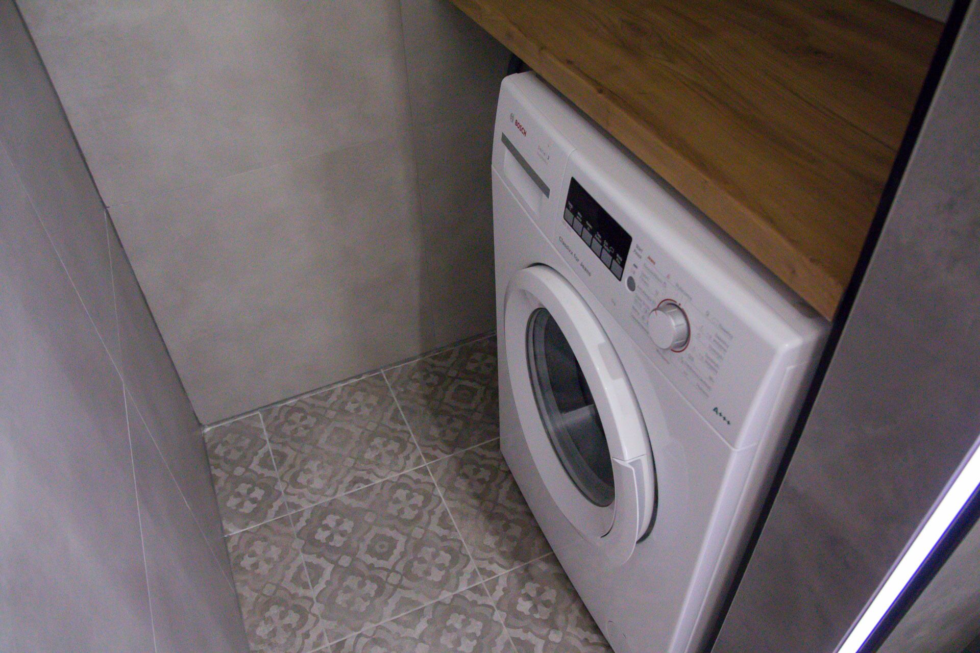 A front-loading washing machine in a small laundry room with patterned tiles and a wooden countertop above it.