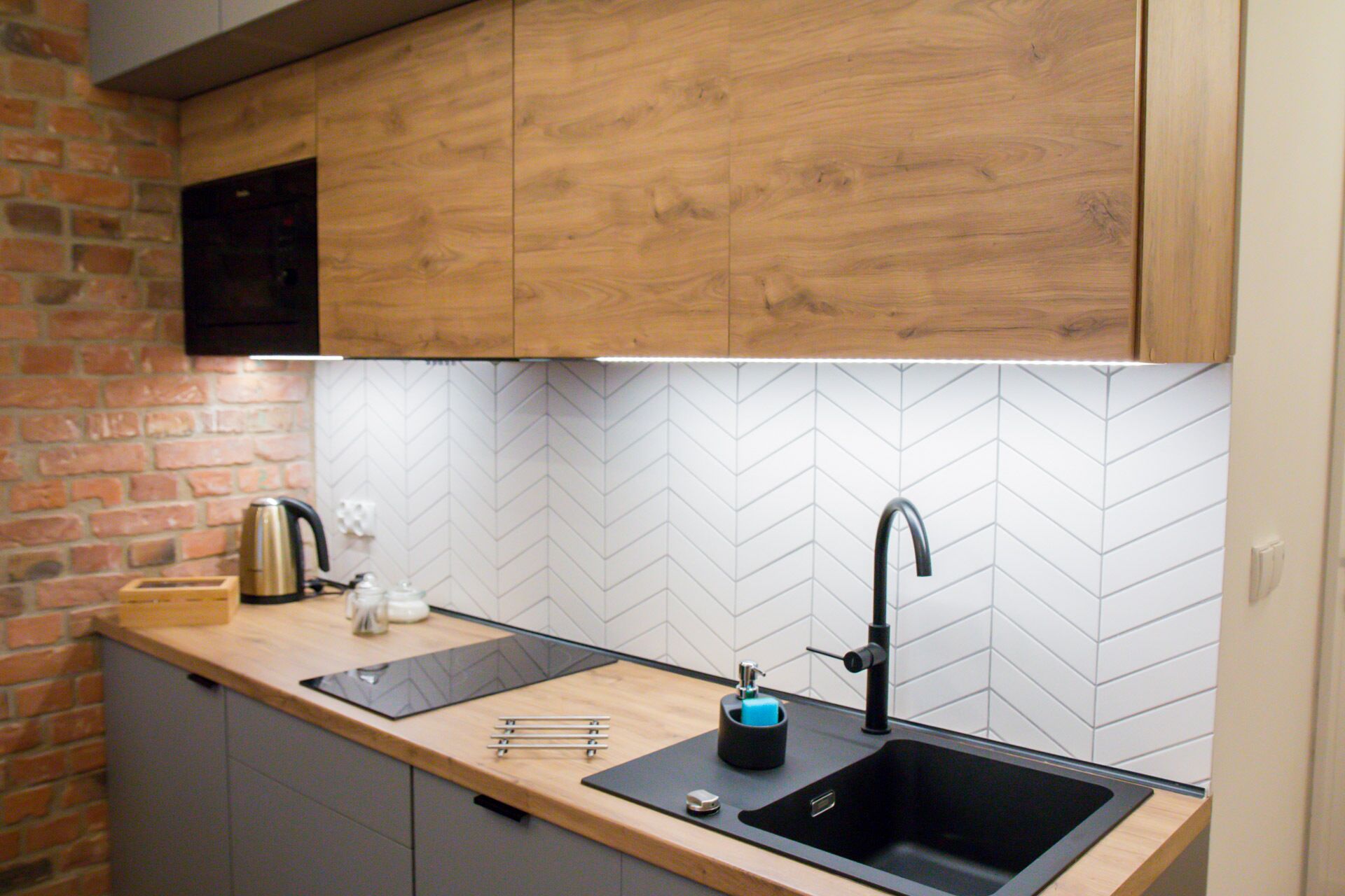 A modern kitchen with a black sink, wooden cabinets, and a herringbone tile backsplash.