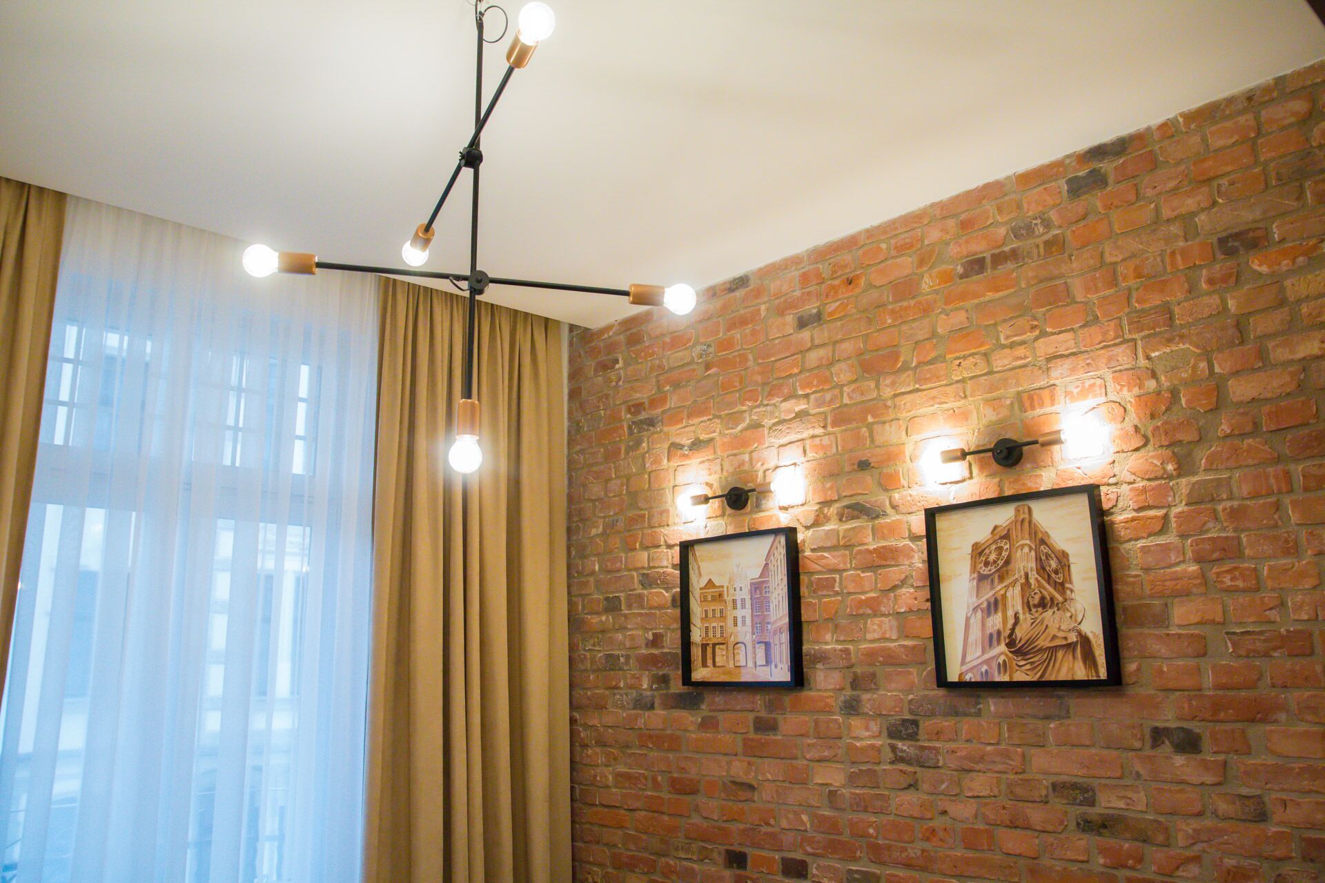 A room with a brick wall, two framed pictures, a hanging light fixture, and a window with curtains.