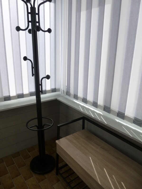 Esquina de una habitación con un banco de madera, un perchero negro y una ventana con persianas verticales.