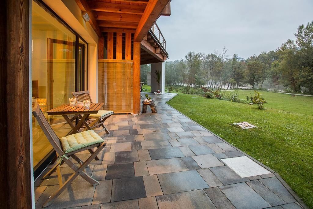 A covered patio with a wooden table and chairs, a stone pathway, and a view of a grassy area with trees.