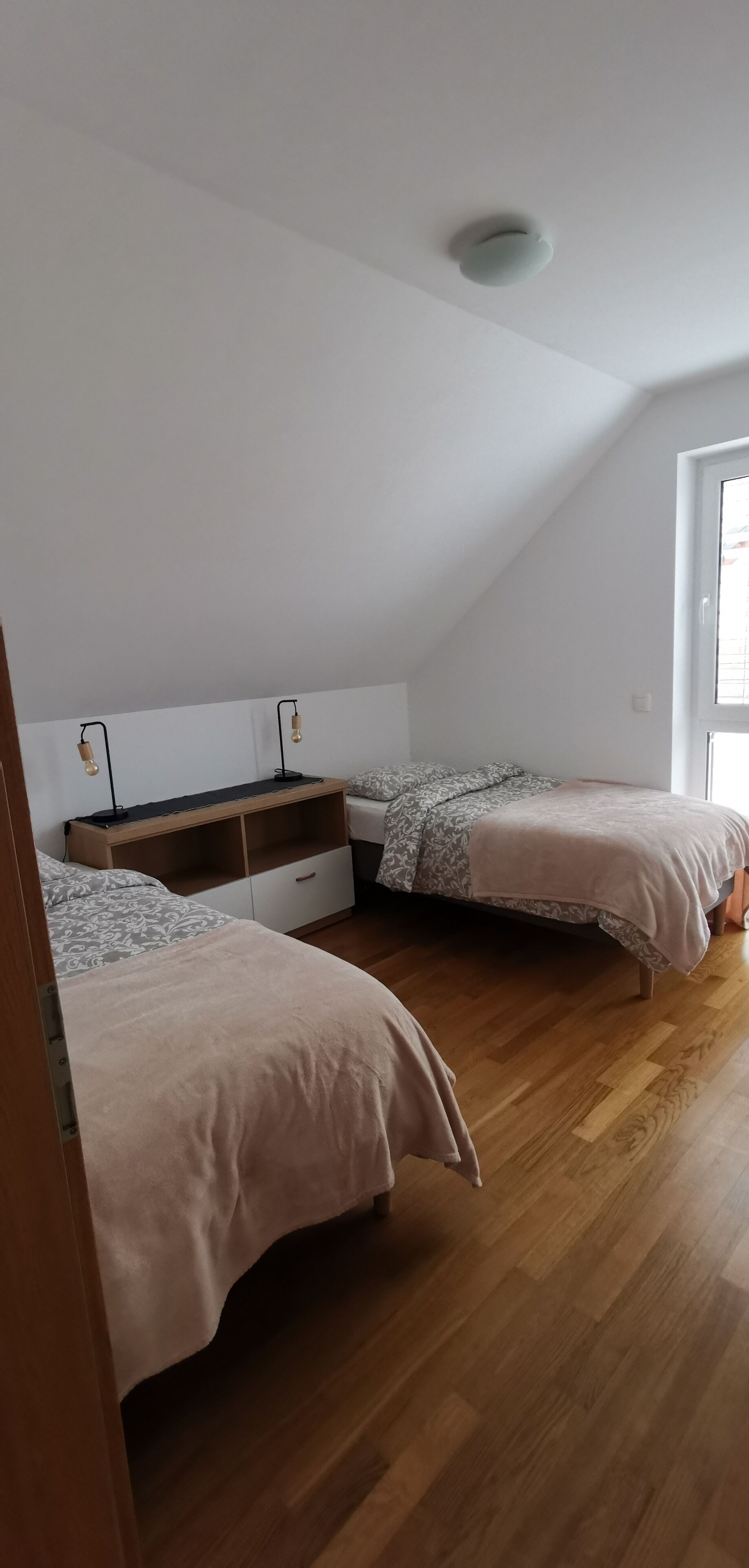 Een slaapkamer met twee bedden, een houten vloer, een wit plafond en een raam.