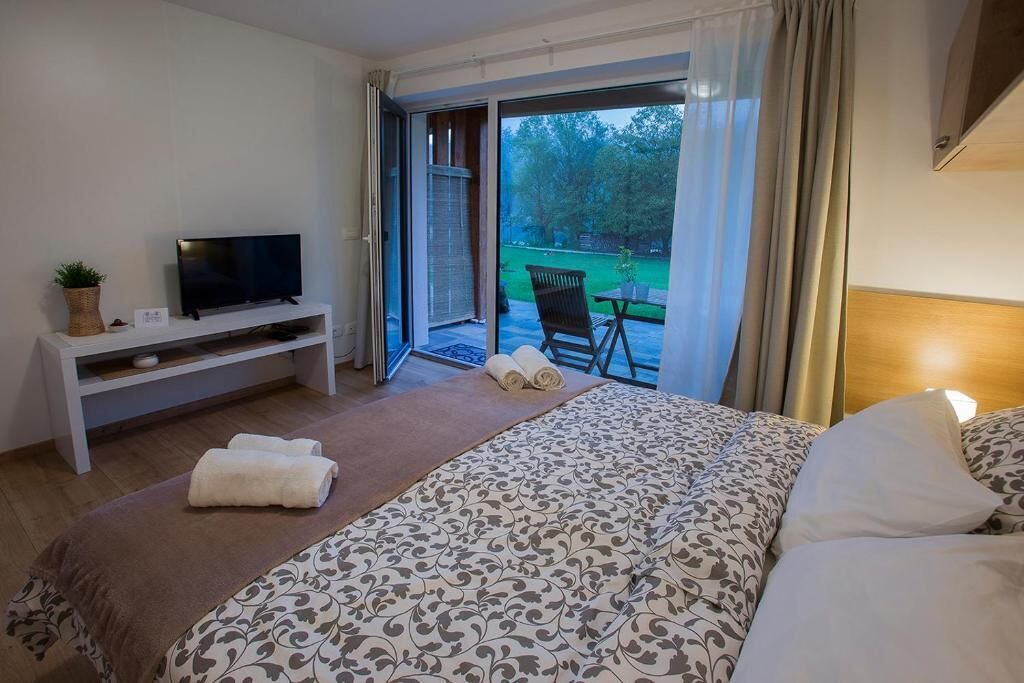 Ein Hotelzimmer mit Bett, Fernseher auf einem Schrank, einem kleinen Tisch mit Stuhl und Blick ins Freie durch eine Schiebetür.