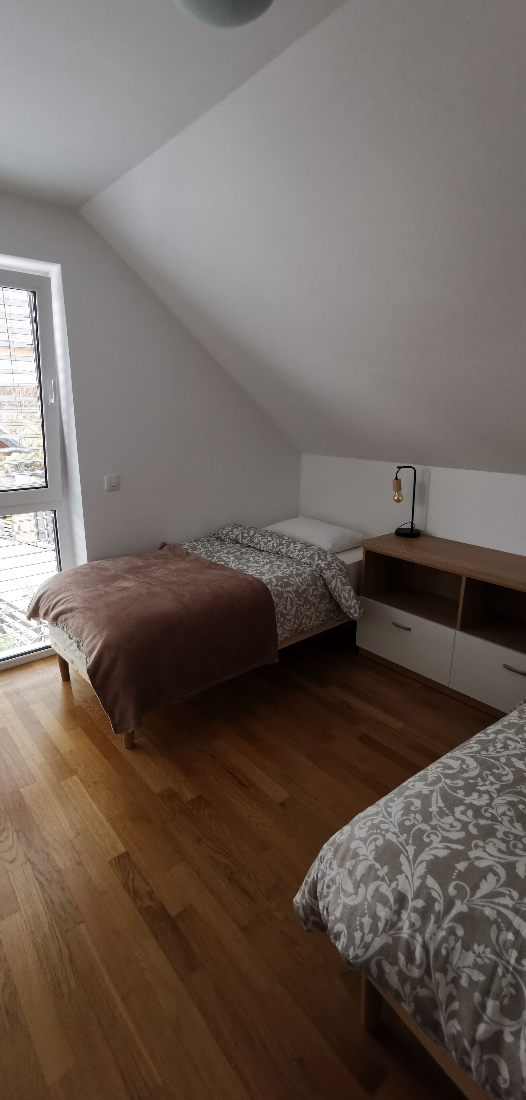 A small, well-lit room with a sloped ceiling, a bed with patterned bedding, a bedside table with a lamp, and a window providing natural light.