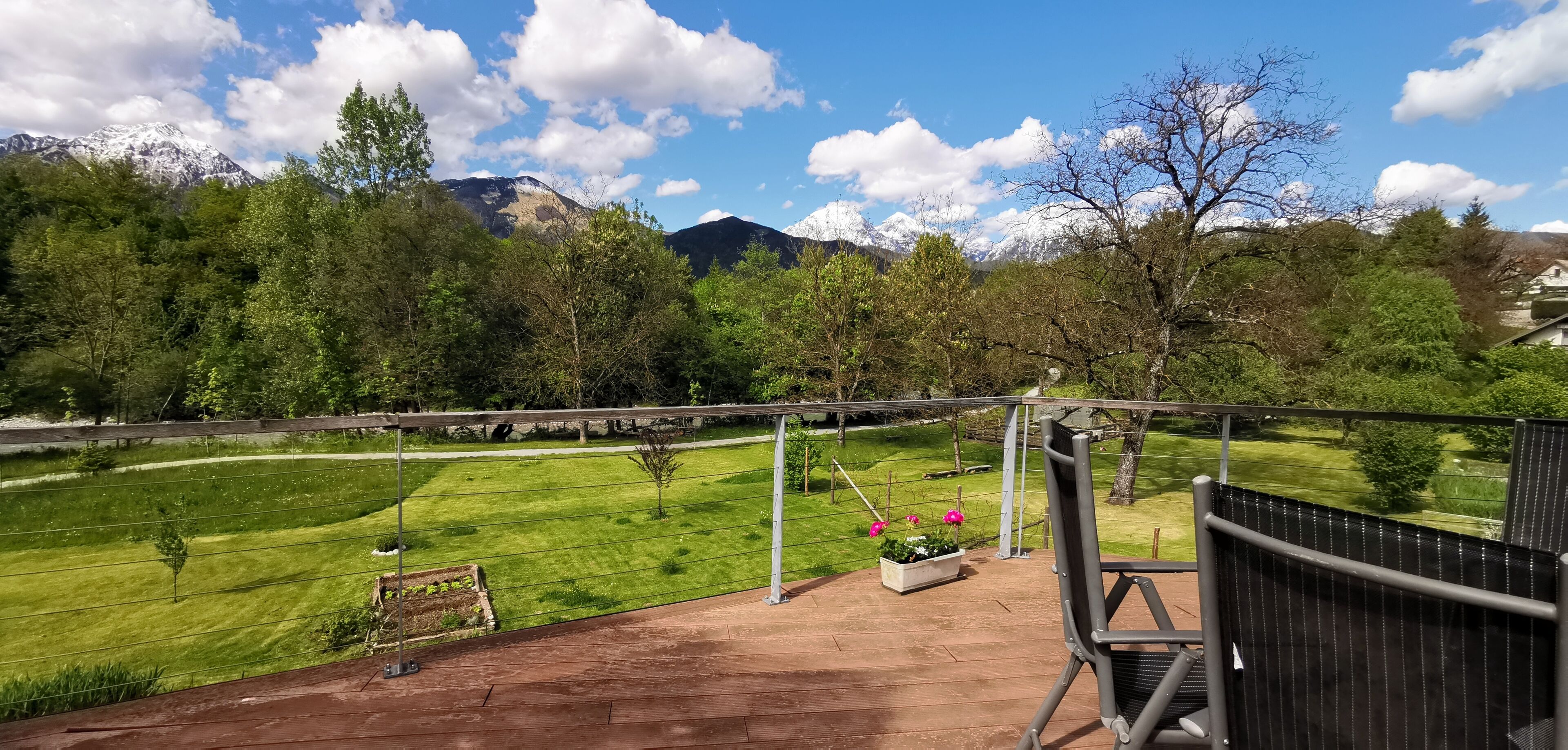 A balcony with a view of a green lawn, a wooden deck, and a mountain range in the background.