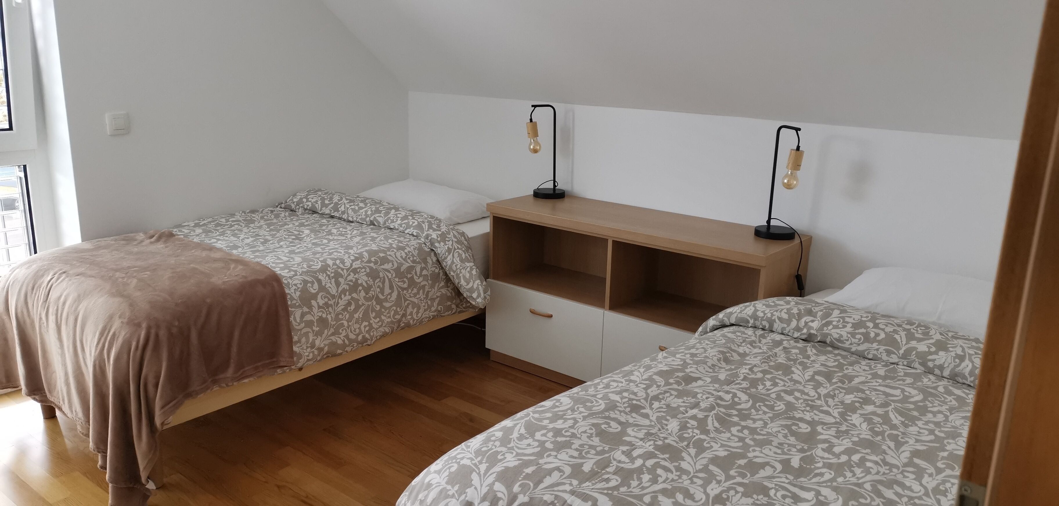A bedroom with two single beds, a wooden nightstand, and two wall-mounted reading lamps.