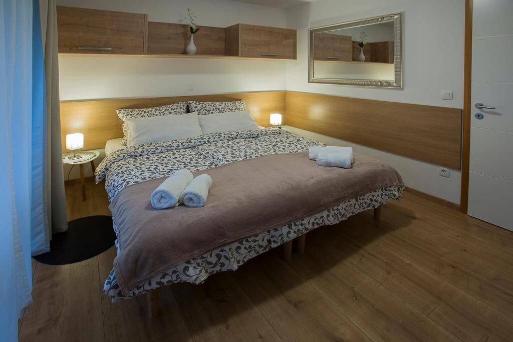 A neatly made bed with a patterned bedspread and two rolled towels, a bedside lamp, and a mirror on the wall.