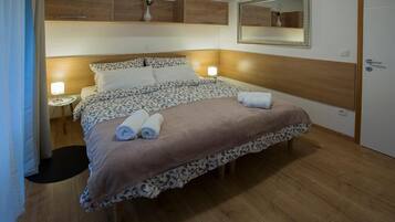 A neatly made bed with a patterned bedspread and two rolled towels, a bedside lamp, and a mirror on the wall.