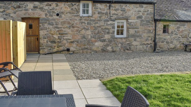 A stone building with a wooden door and windows, a gravel driveway, and outdoor seating with chairs and a table. - 2 bedroom accommodation in Morfa Nefyn (Morfa Nefyn)