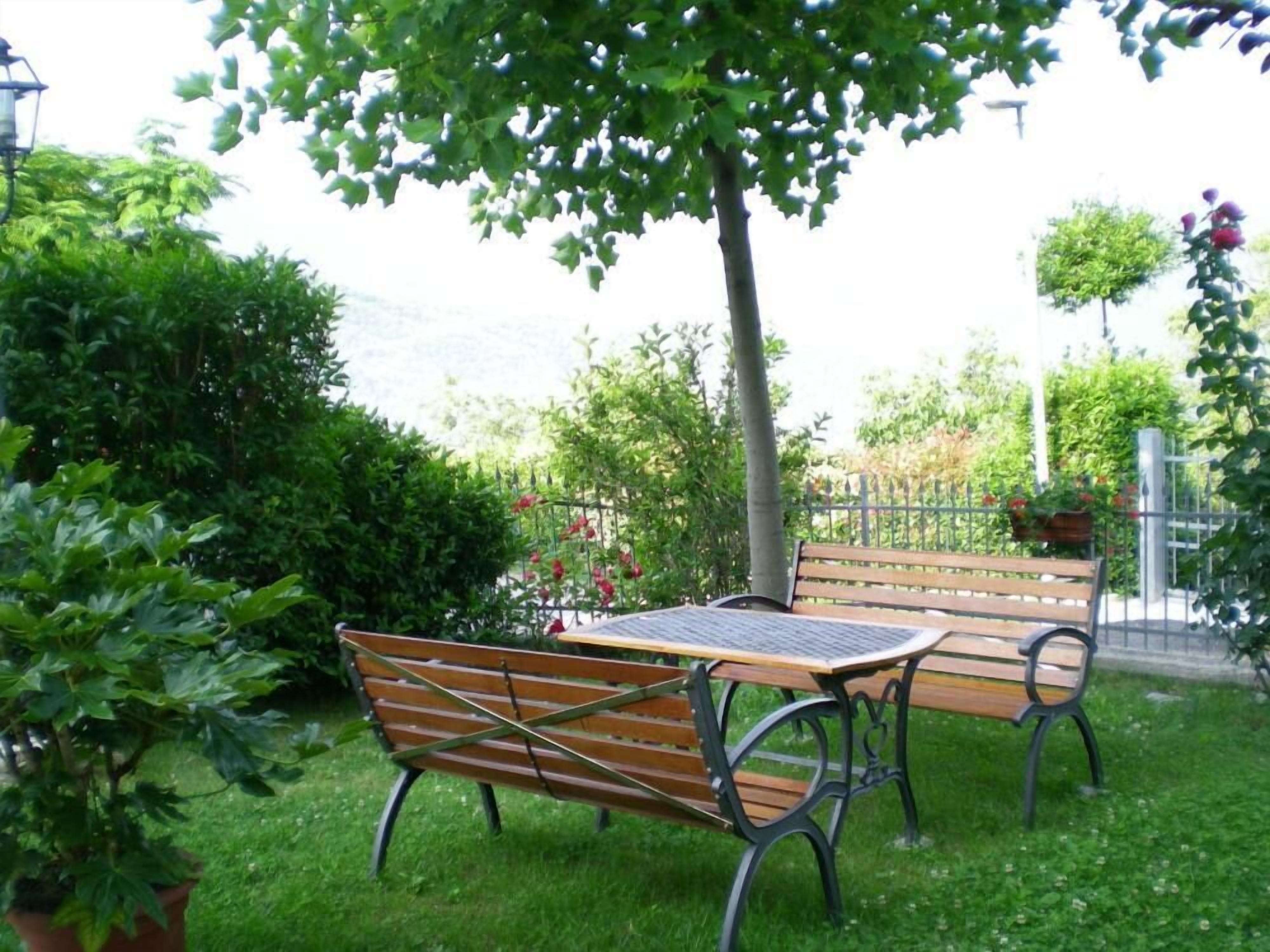 A bench and table set in a lush garden with a metal fence and greenery in the background.