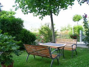 Ein Bank- und Tischset in einem üppigen Garten mit einem Metallzaun und Grünpflanzen im Hintergrund.