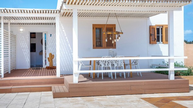 En modern uteplats med en vit pergola, träpersienner och en matplats med vita stolar och ett bord.