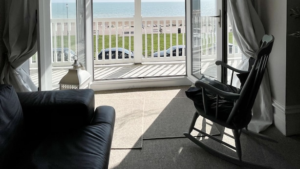 A room with a large bay window overlooking a grassy area and the sea. There is a black leather sofa and a rocking chair. - Stunning Sea Front Apartment (Hythe)