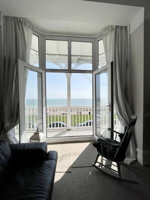 A room with a large bay window overlooking a grassy area and the sea. There is a black leather sofa and a rocking chair. - Stunning Sea Front Apartment (Hythe)