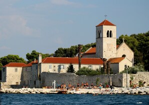 A historic stone building with a red-tiled roof, a bell tower, and a waterfront setting with people swimming and sunbathing. - C - apt w. Balcony, Shared Terrace & the sea View (Trogir)