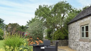 Un espacio al aire libre con mobiliario de mimbre, rodeado de un muro de piedra y un jardín con diversas plantas y flores.