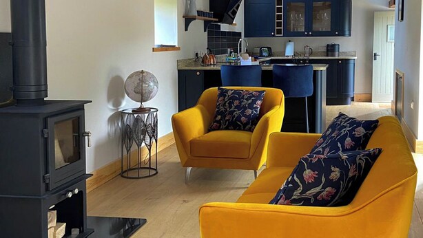 Een moderne woonkamer met een zwart houthakkerkacheltje, twee gele fauteuils met bloemenkussens, een klein bijzettafeltje met een wereldbol, en een keuken op de achtergrond.