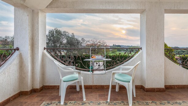 Un balcón con dos sillas blancas y una mesa, con vistas a un jardín y una puesta de sol.